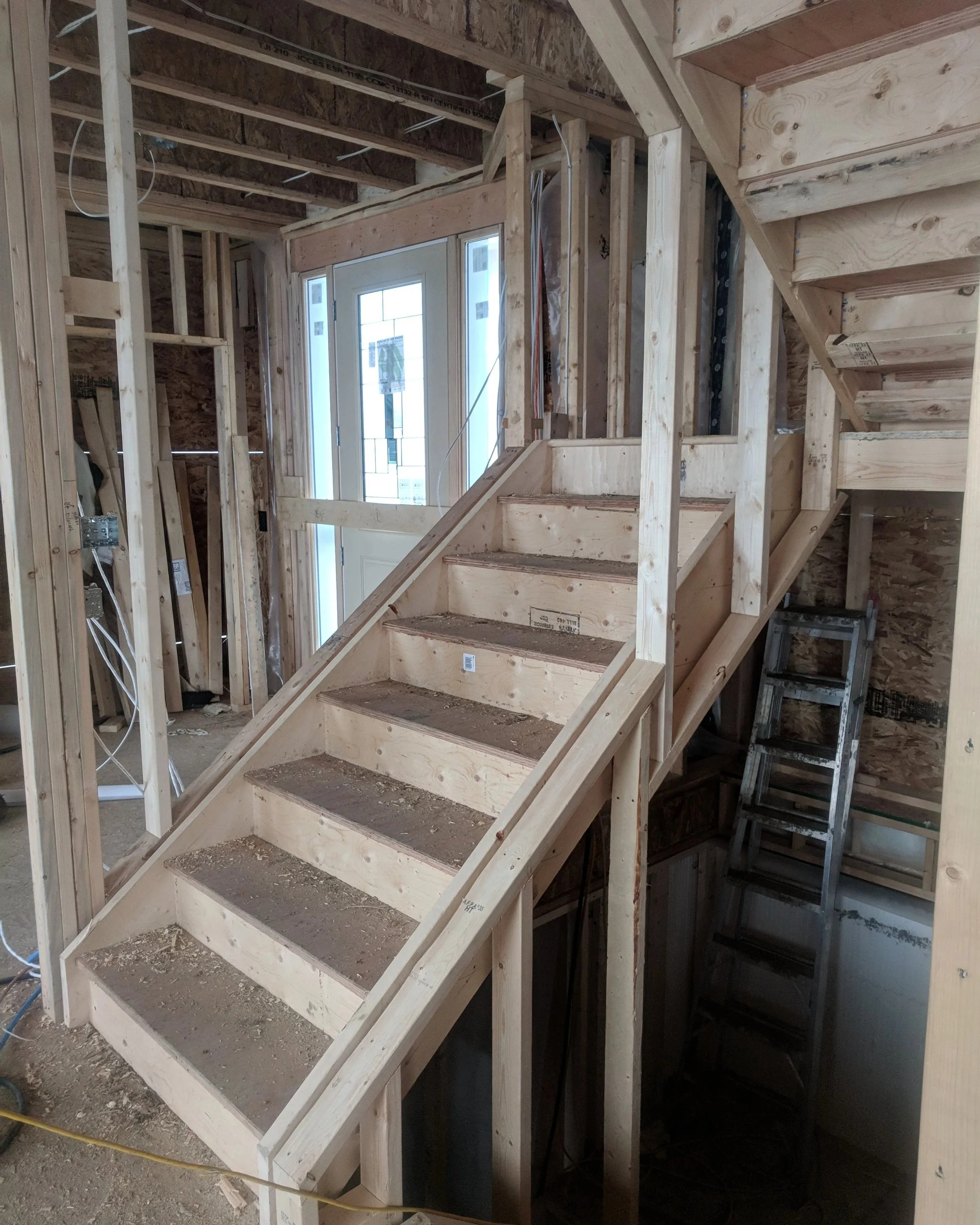 Wooden staircase under construction inside a house with exposed framing, electrical wiring, and a front door with glass panes.
