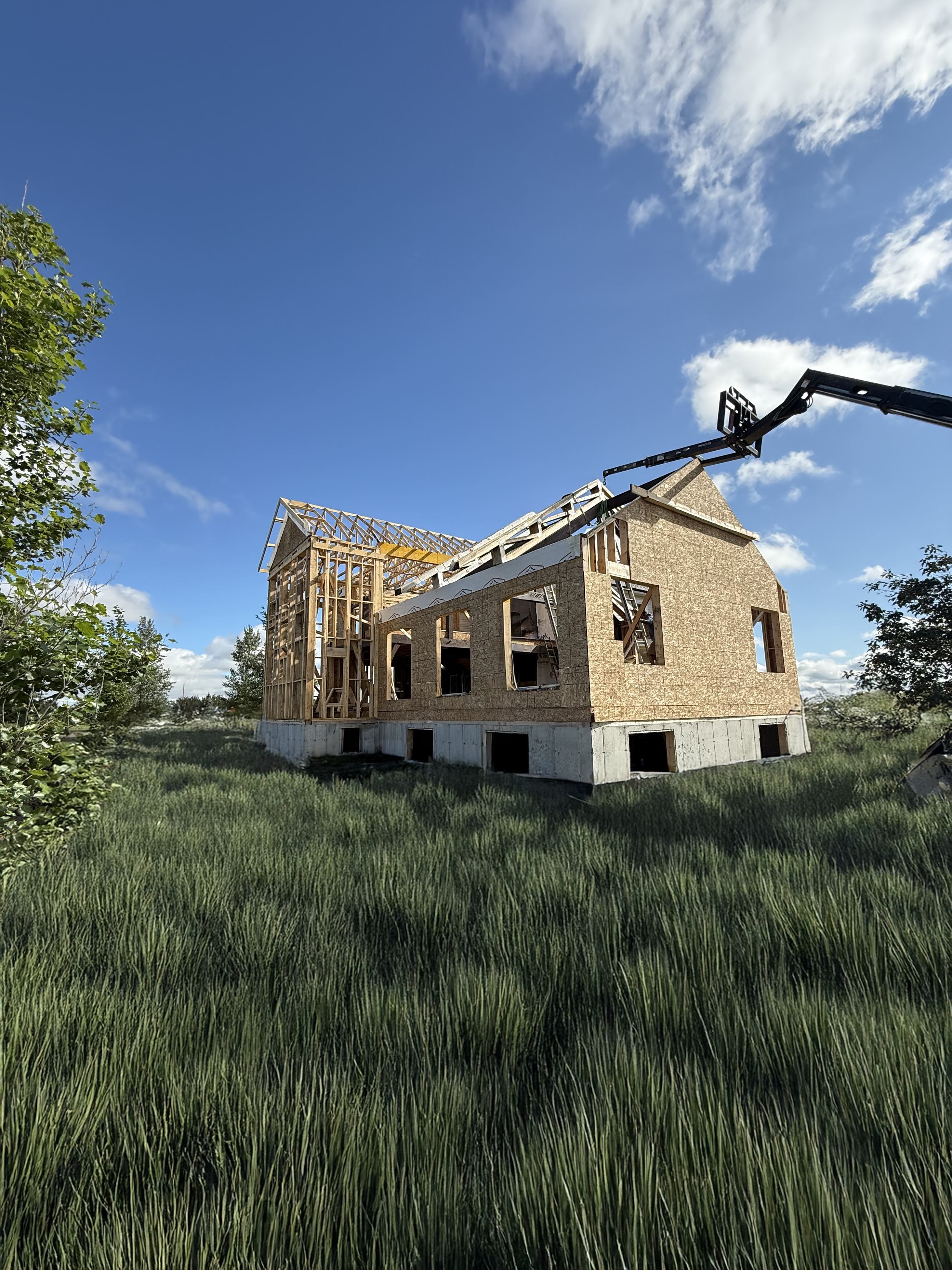 Construction of a house with wooden framing and a concrete foundation, surrounded by green grass and trees, under a blue sky with scattered clouds.