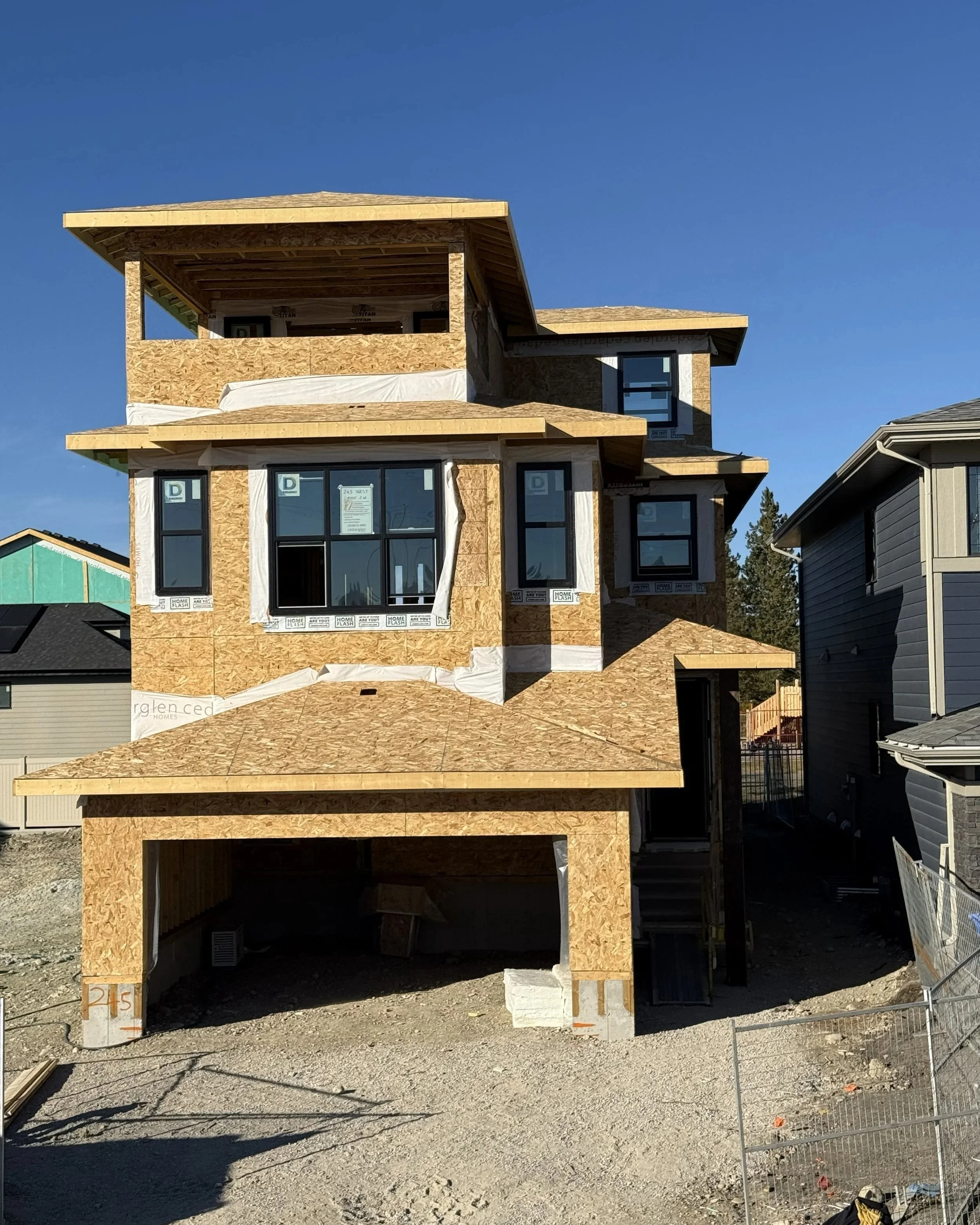 A multi-story house under construction with plywood siding and large windows, next to an unfinished driveway and neighboring houses.
