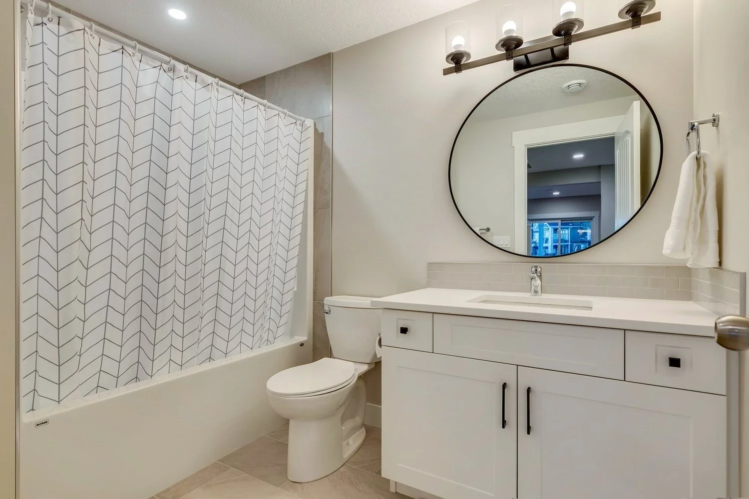 Bathroom with a bathtub and shower with patterned curtain, a toilet, and a white vanity with a round mirror and a wall-mounted light fixture.
