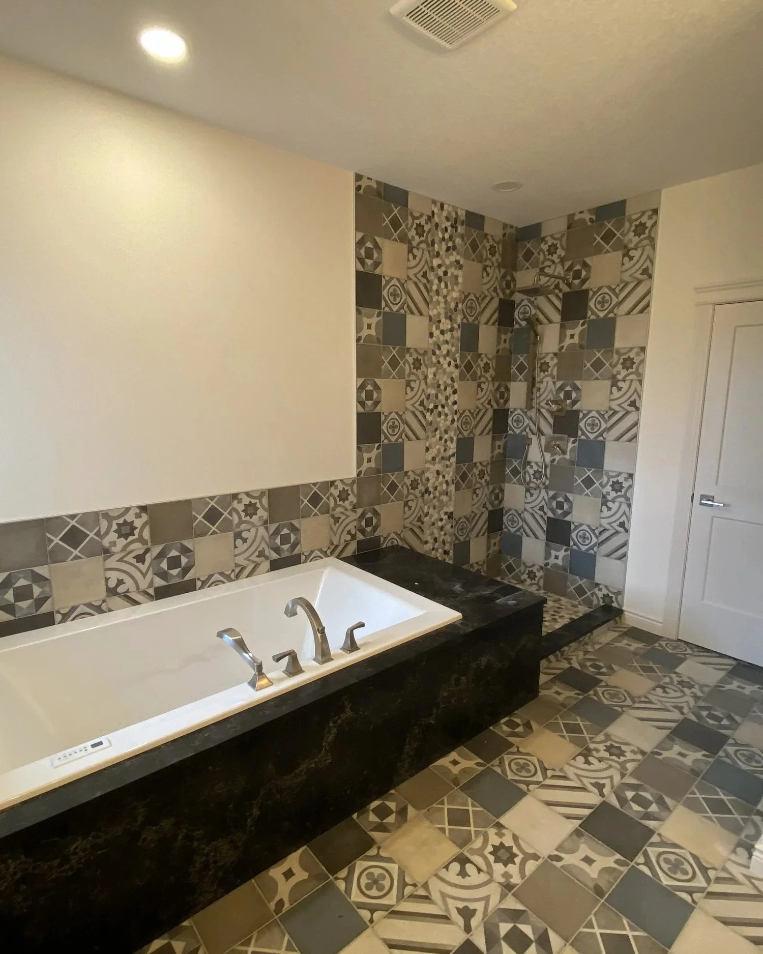 A modern bathroom with a built-in bathtub surrounded by black marble. The wall behind the tub and the walk-in shower area are decorated with patterned tiles in shades of gray, black, and beige. The bathroom has a white ceiling with recessed lighting and an exhaust vent, and a white door.