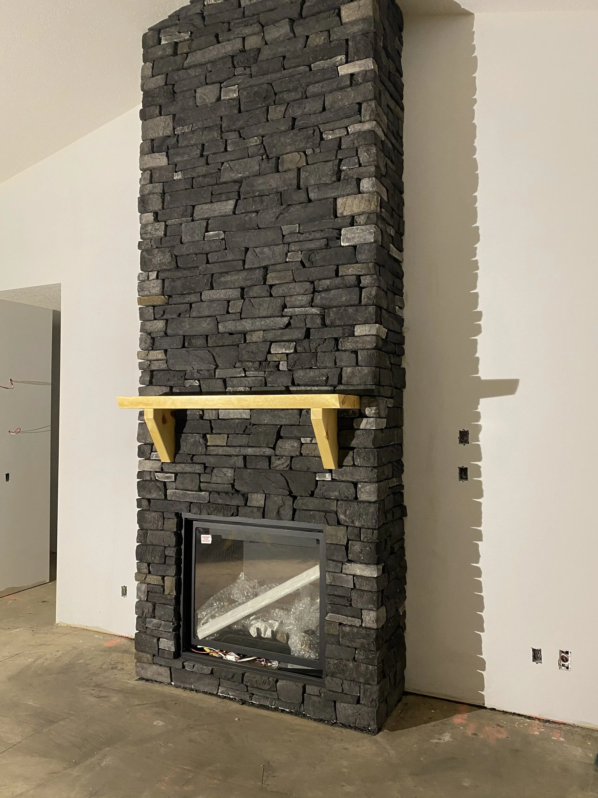 A black stone fireplace with a glass door at the bottom and a wooden mantel shelf above it, under construction or renovation.