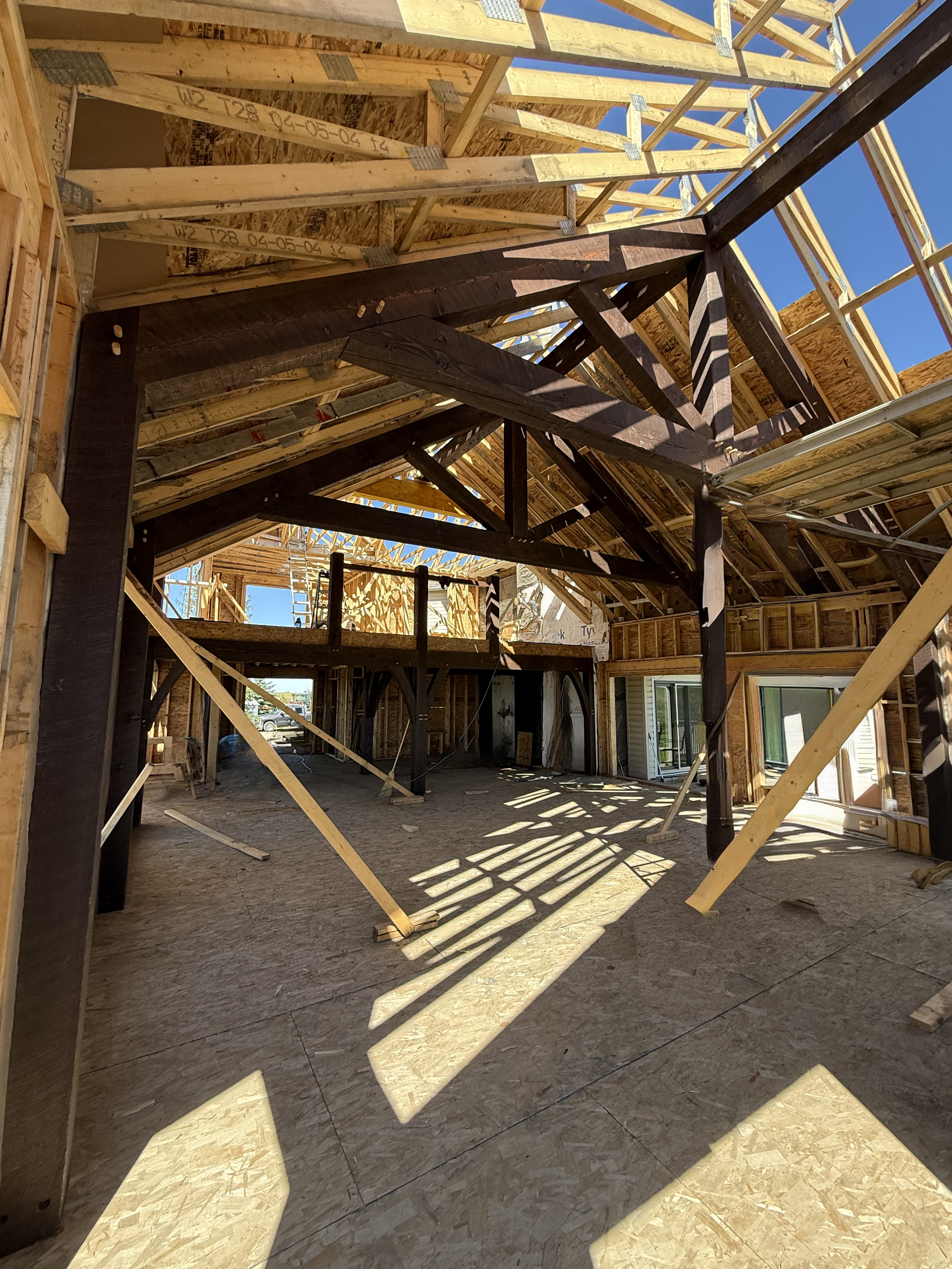 Interior of a house under construction with exposed wooden framing and ceiling rafters, sunlight casting shadows on the plywood subfloor.