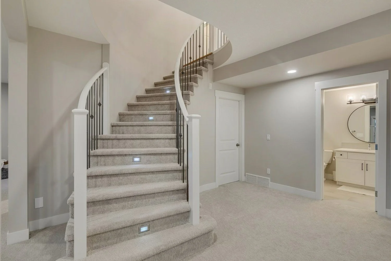 Interior view of a modern home with a curved staircase carpeted in beige, leading to an upper floor, and a visible bathroom with a vanity mirror and toilet.