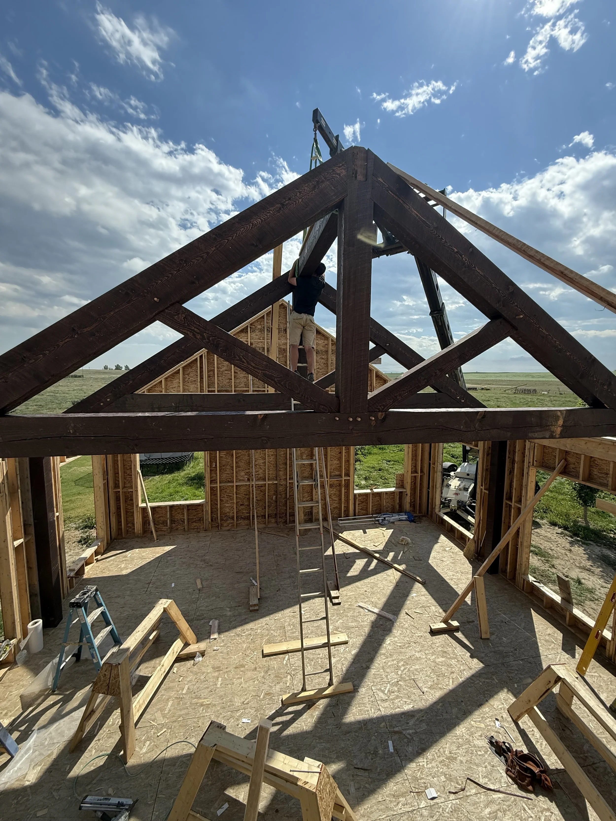 Construction of a wooden house frame with a man working on the roof trusses outdoors under a cloudy sky.
