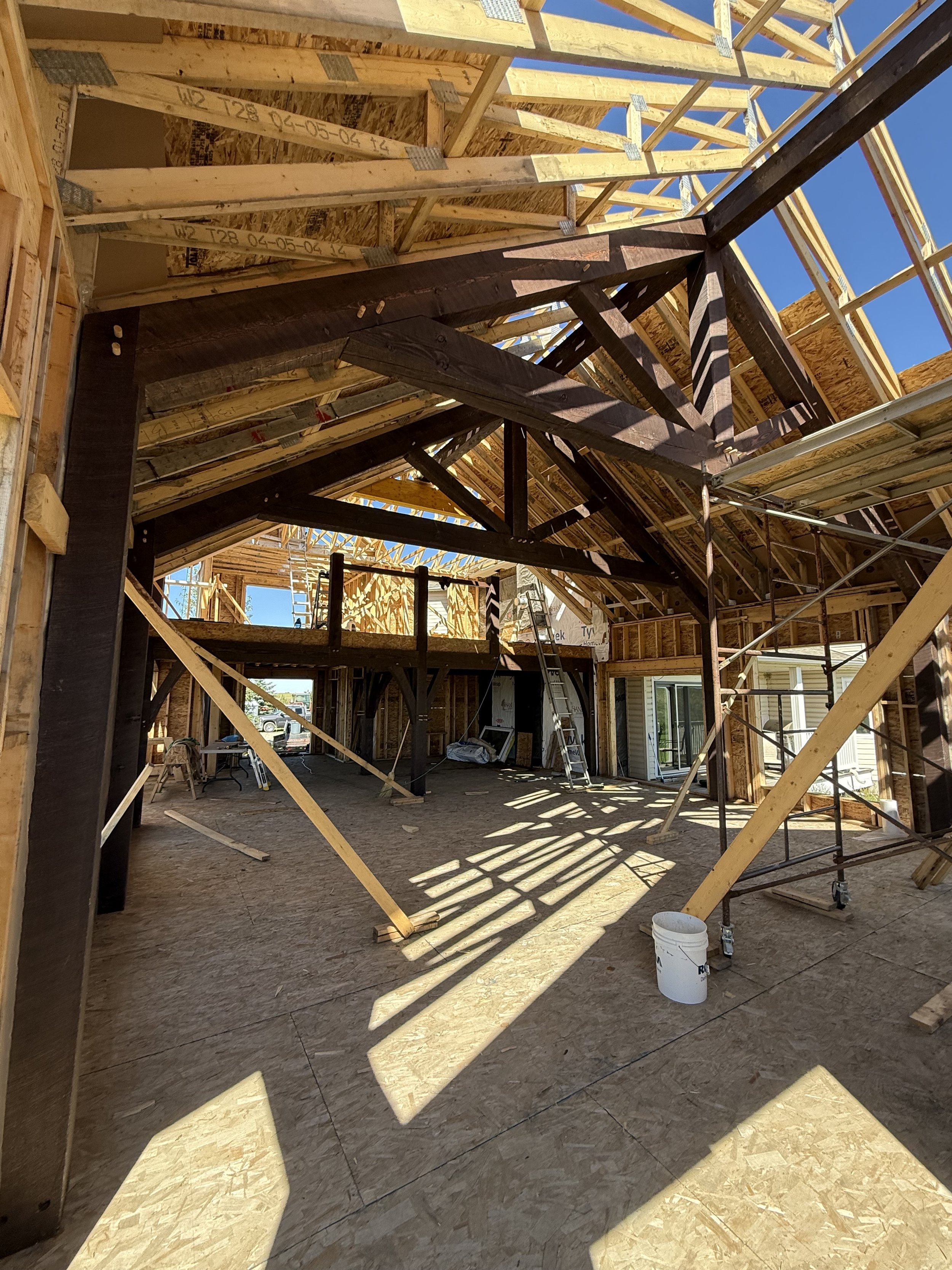 Interior of a house under construction with wooden framing, scaffolding, and plywood floors, sunlight casting shadows through open walls.