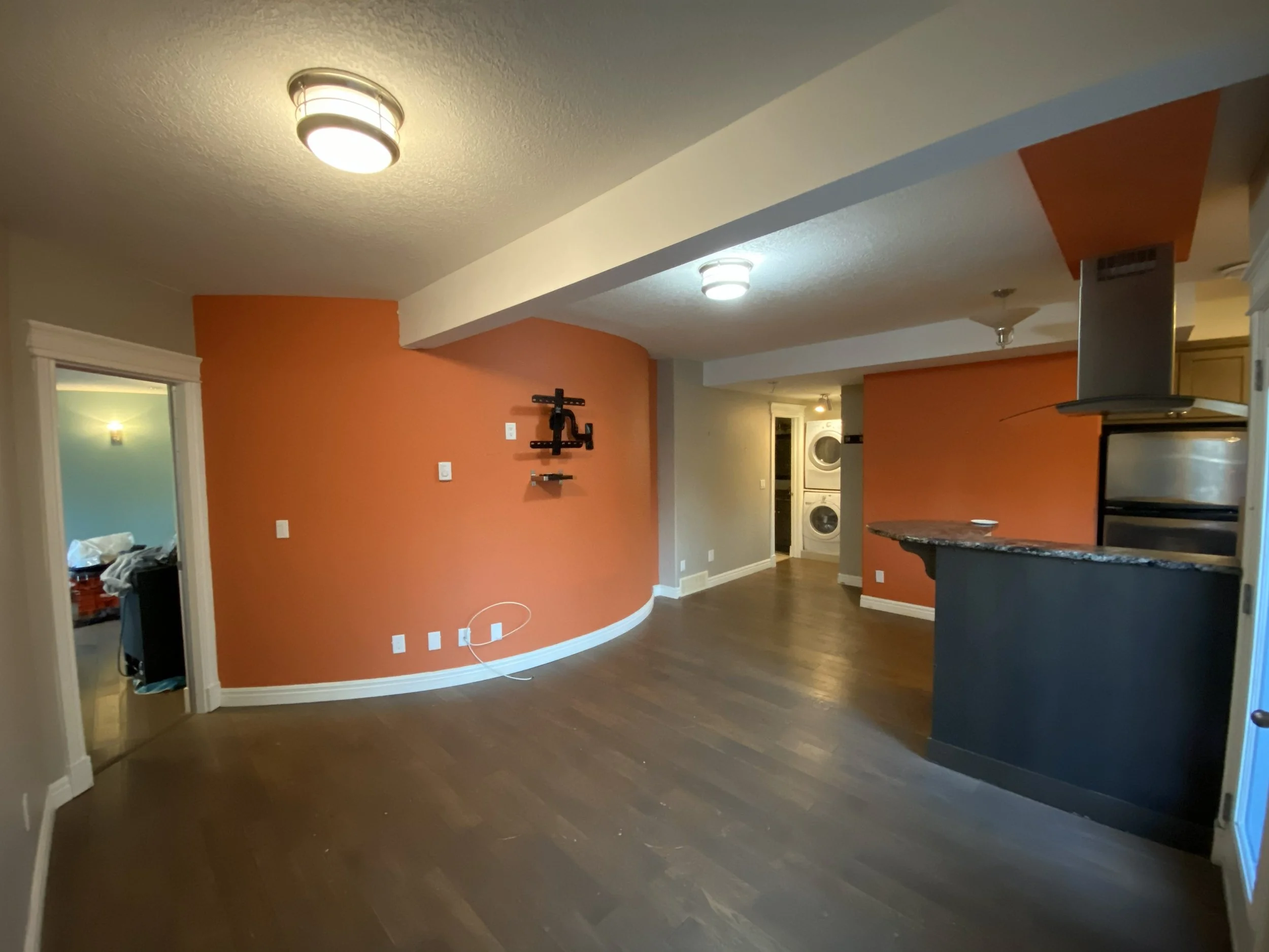 Empty living room with hardwood floors, orange accent walls, and ceiling lights. A doorway leads to a cluttered bedroom, and a laundry area with stacked washers and dryers is in the background.