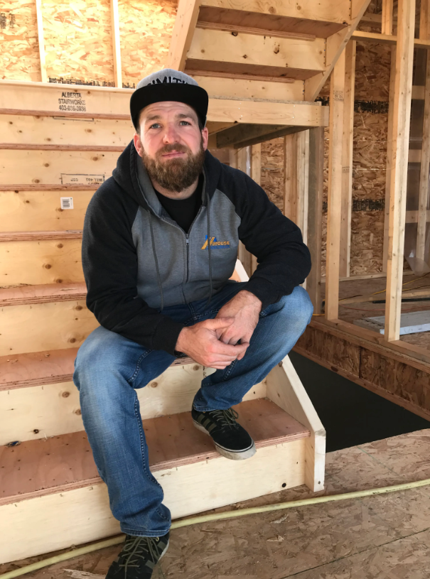 Man sitting on wooden stairs inside a construction site, wearing a gray and black hoodie, blue jeans, black sneakers, and a baseball cap, with unfinished wooden framing around him.