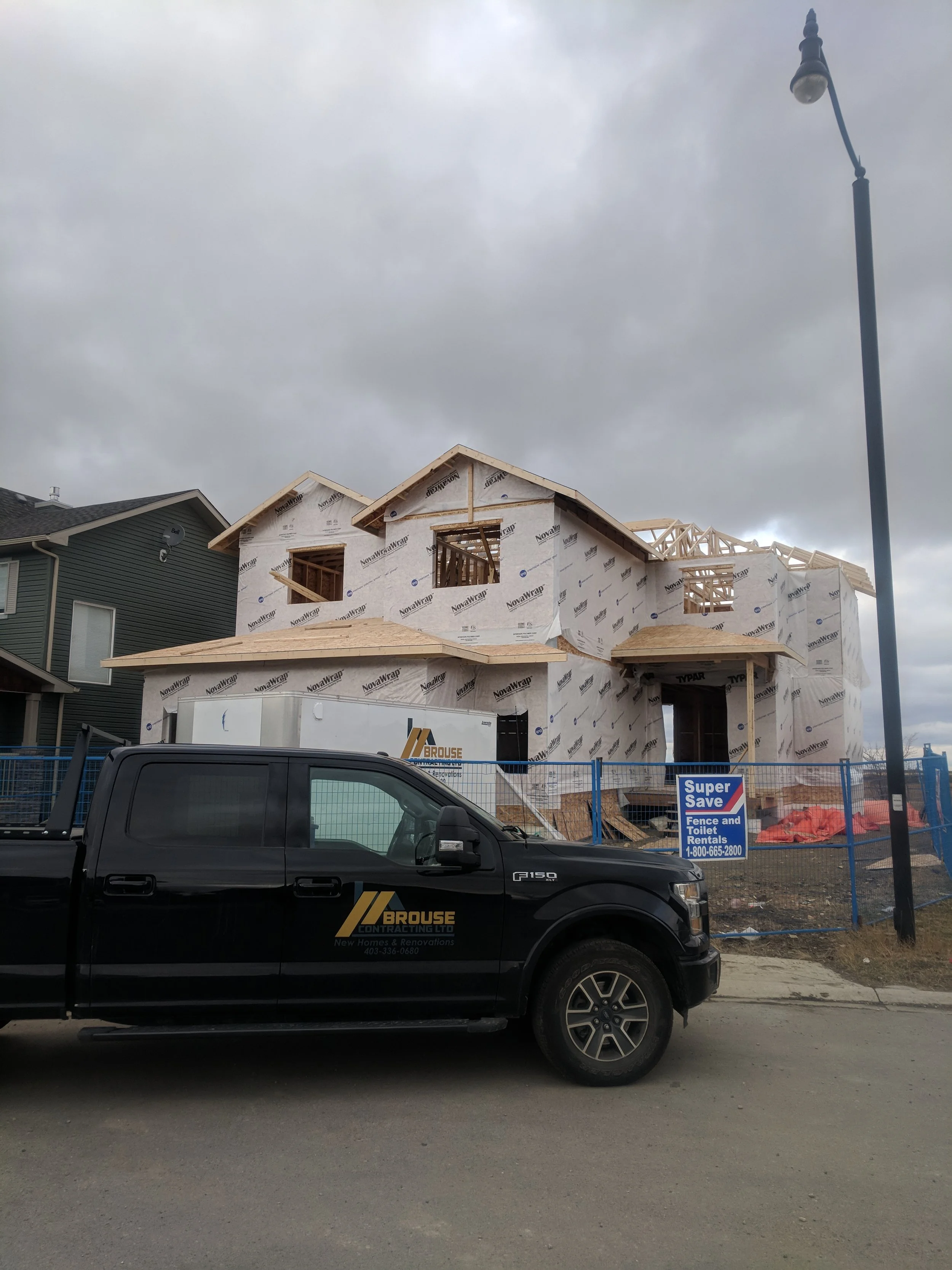 A two-story house under construction with wrapped exterior walls, wooden framing visible around windows and roof, and construction vehicles parked in front. Overcast sky, residential neighborhood, and street lamp in the scene.