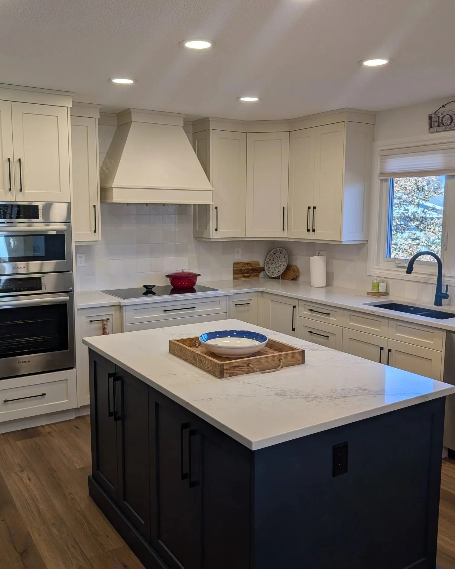Modern kitchen with white cabinetry, black hardware, and a central black and white island with marble countertop. There are built-in double ovens, a red pot on the stovetop, and various kitchen decor items. A window with a view of trees is above the sink.