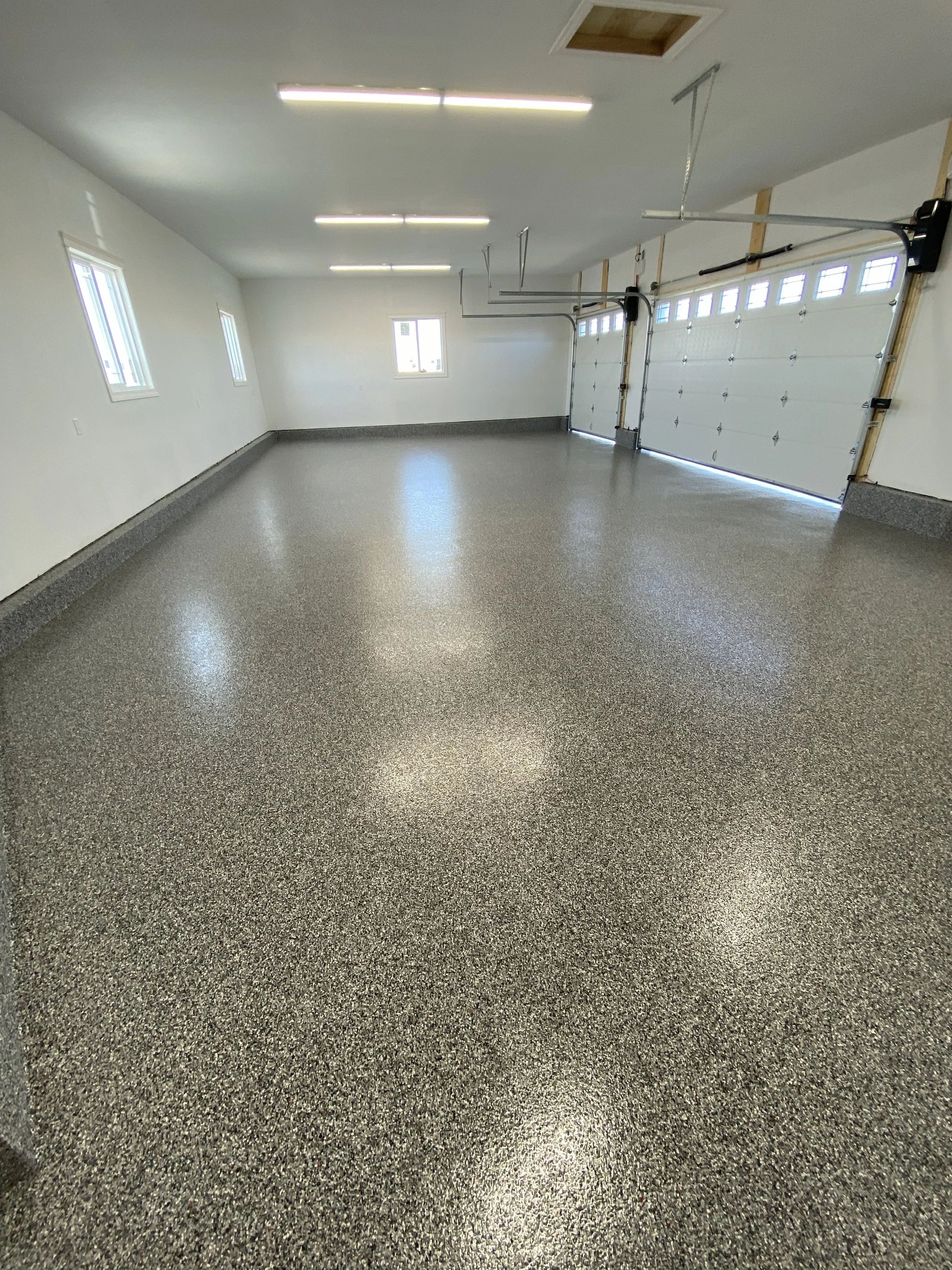 Empty spacious garage with speckled epoxy floor, white walls, three small windows, and two white garage doors.