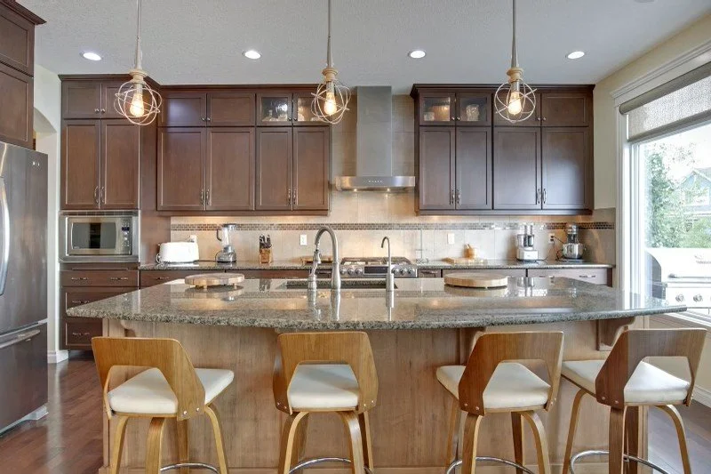 Modern kitchen with brown cabinets, granite island, stainless steel appliances, pendant lights, and barstools.