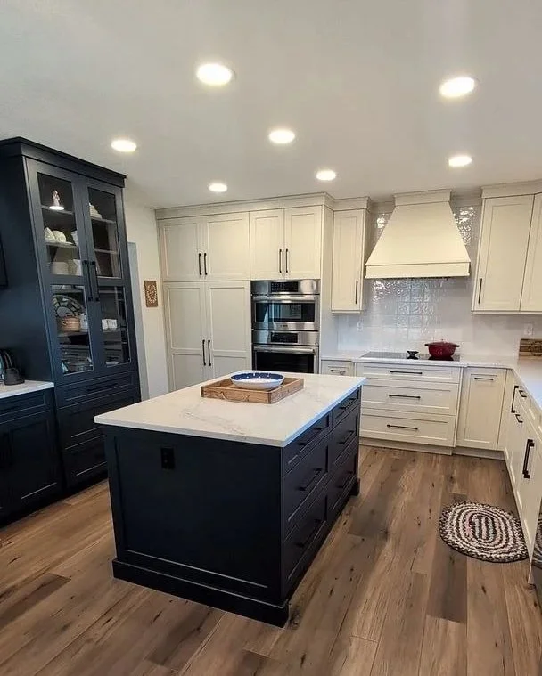 Modern kitchen with white cabinets, a dark island with a white countertop, and a wooden floor. There are built-in ovens, a white range hood, and a blue china cabinet on the left. Recessed ceiling lights illuminate the space.