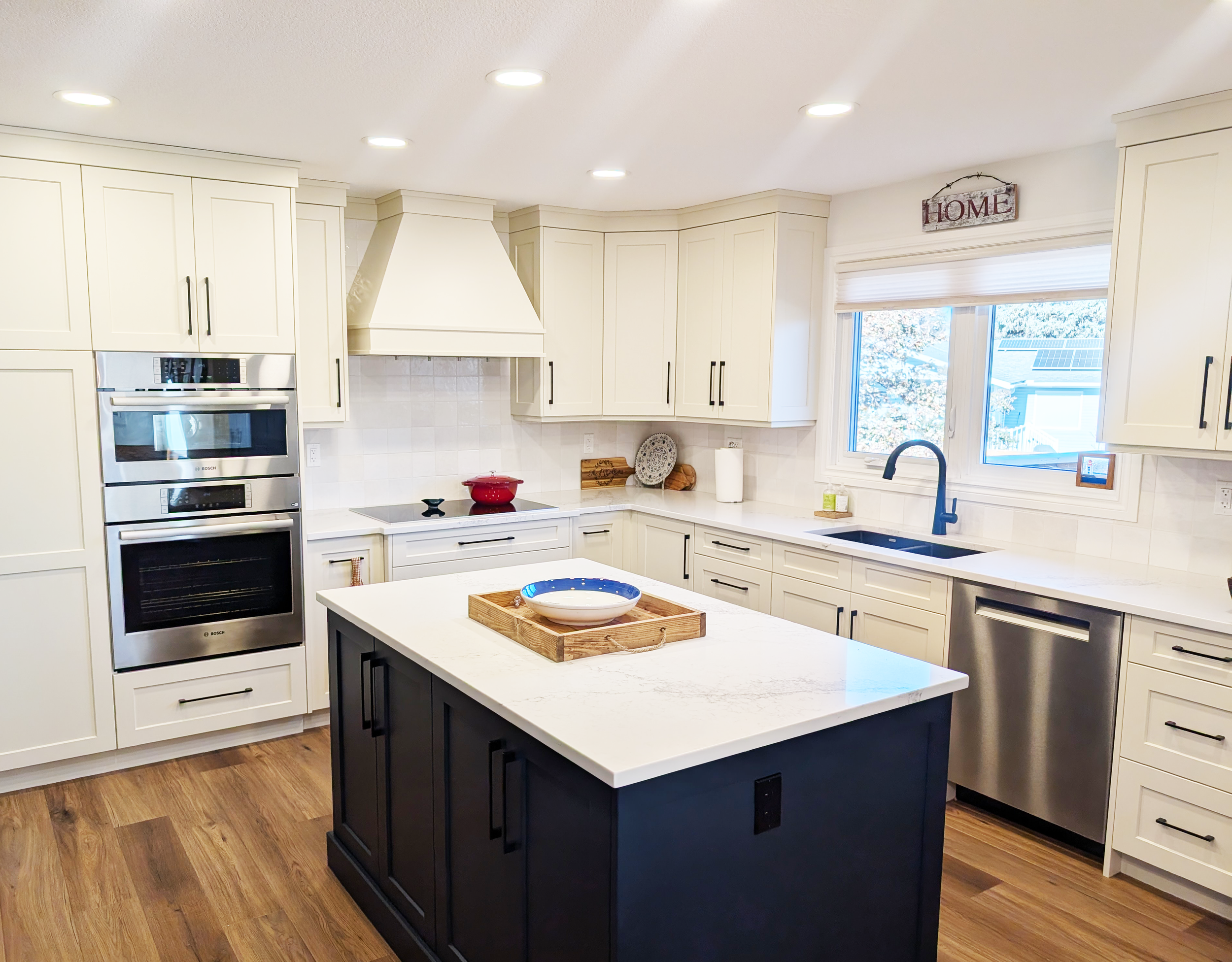 Modern kitchen with white cabinets, a black island, stainless steel appliances, and a window above the sink showing outdoor trees.