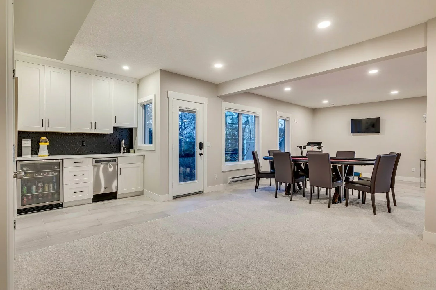 Open-concept living room and kitchen area with white cabinets, black backsplash, dining table with six chairs, a wall-mounted TV, and large windows showing an outdoor view.