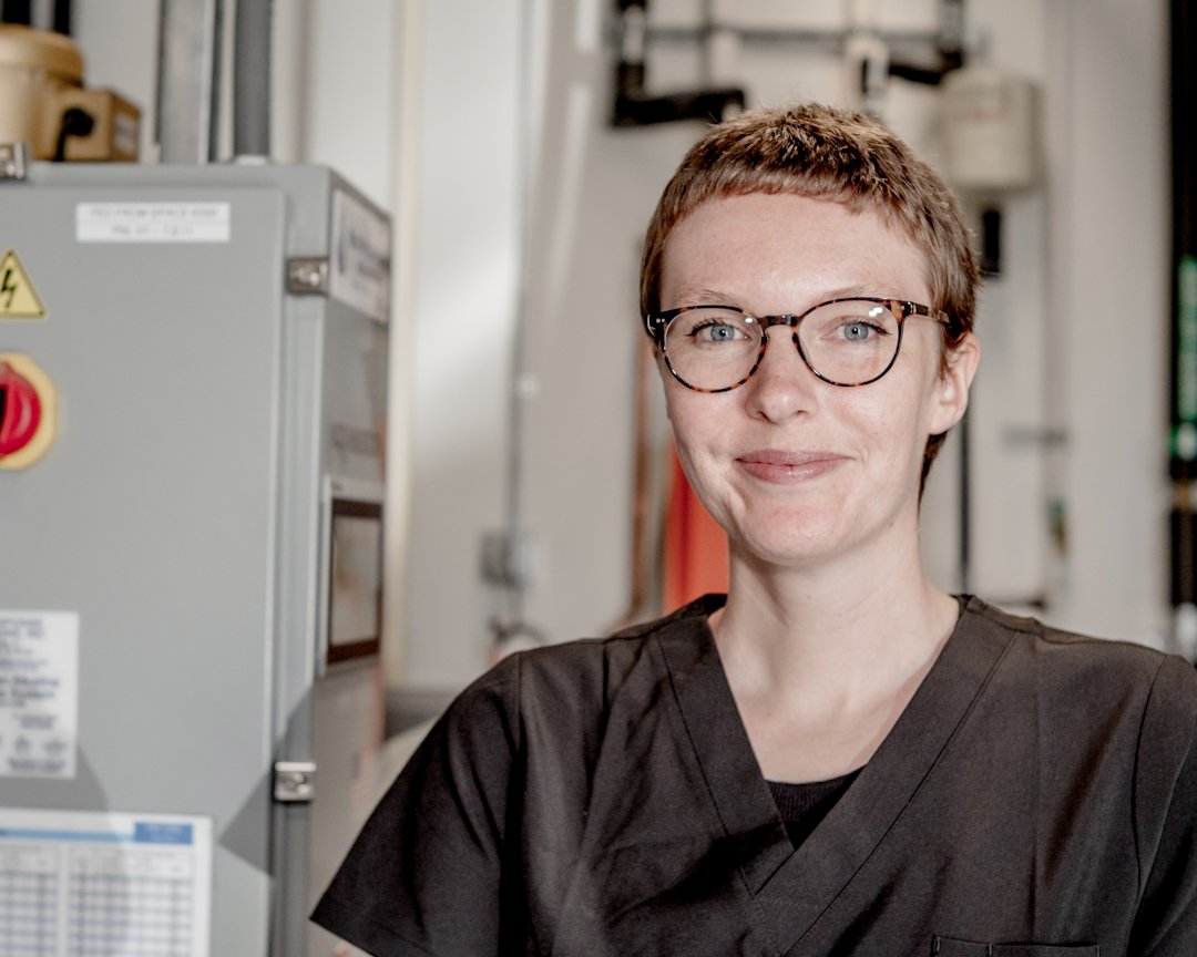 A person in black scrubs with short hair and glasses, smiling in a professional and industrial pet water cremation setting, with equipment in the background.