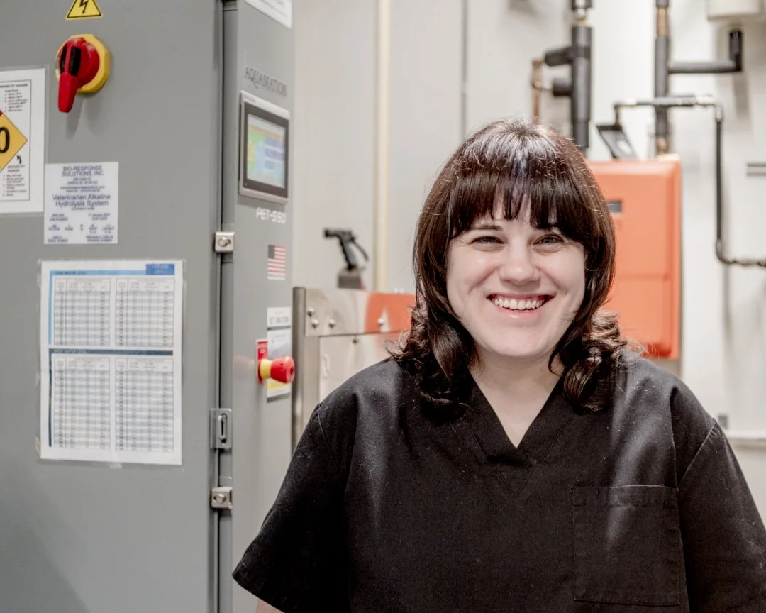 A woman in black scrubs with brown hair and bangs, smiling in a professional and industrial pet water cremation setting, with equipment in the background.
