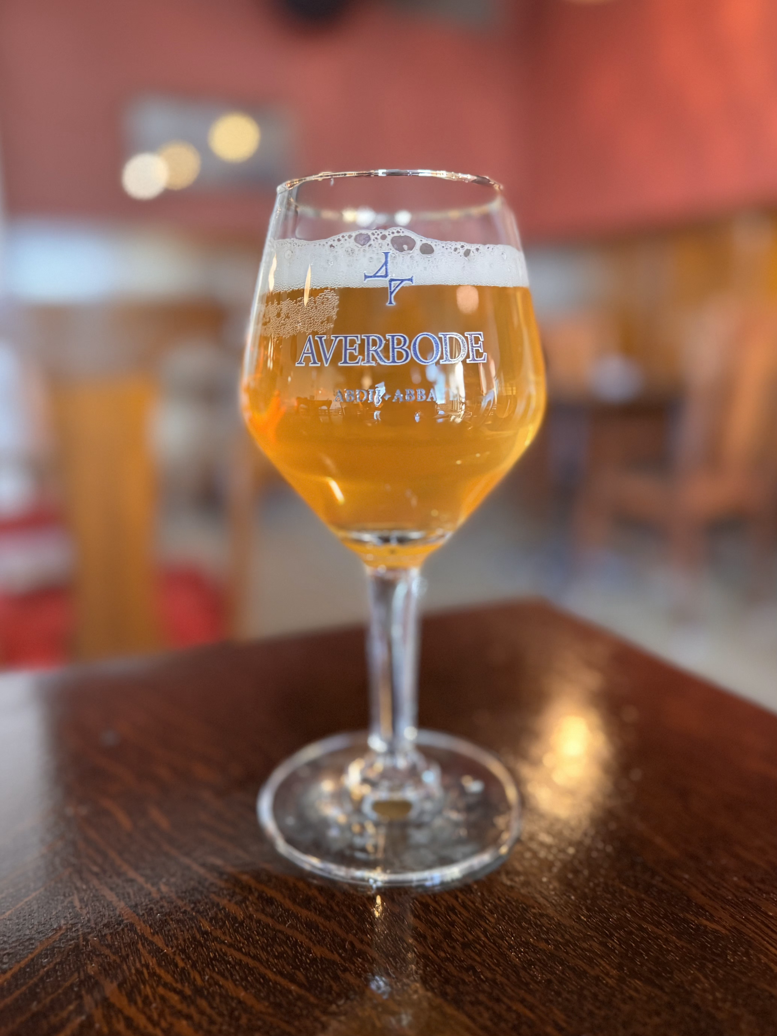A glass of beer on a wooden table with the logo 'AVERBODE' printed on it, with a blurred background of a bar or restaurant setting.