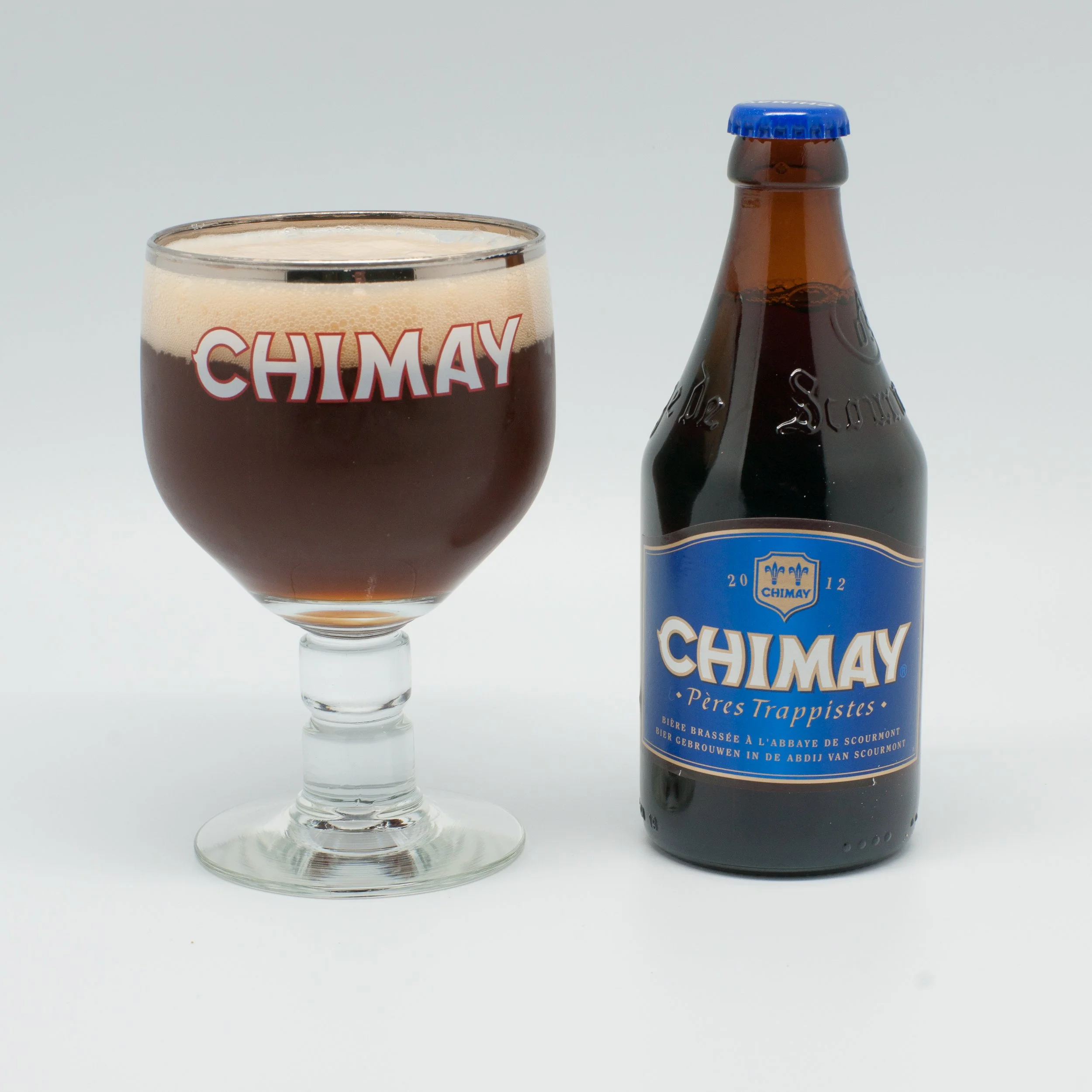 A glass of Chimay beer with a foamy head next to a Chimay beer bottle with a blue label on a white background.