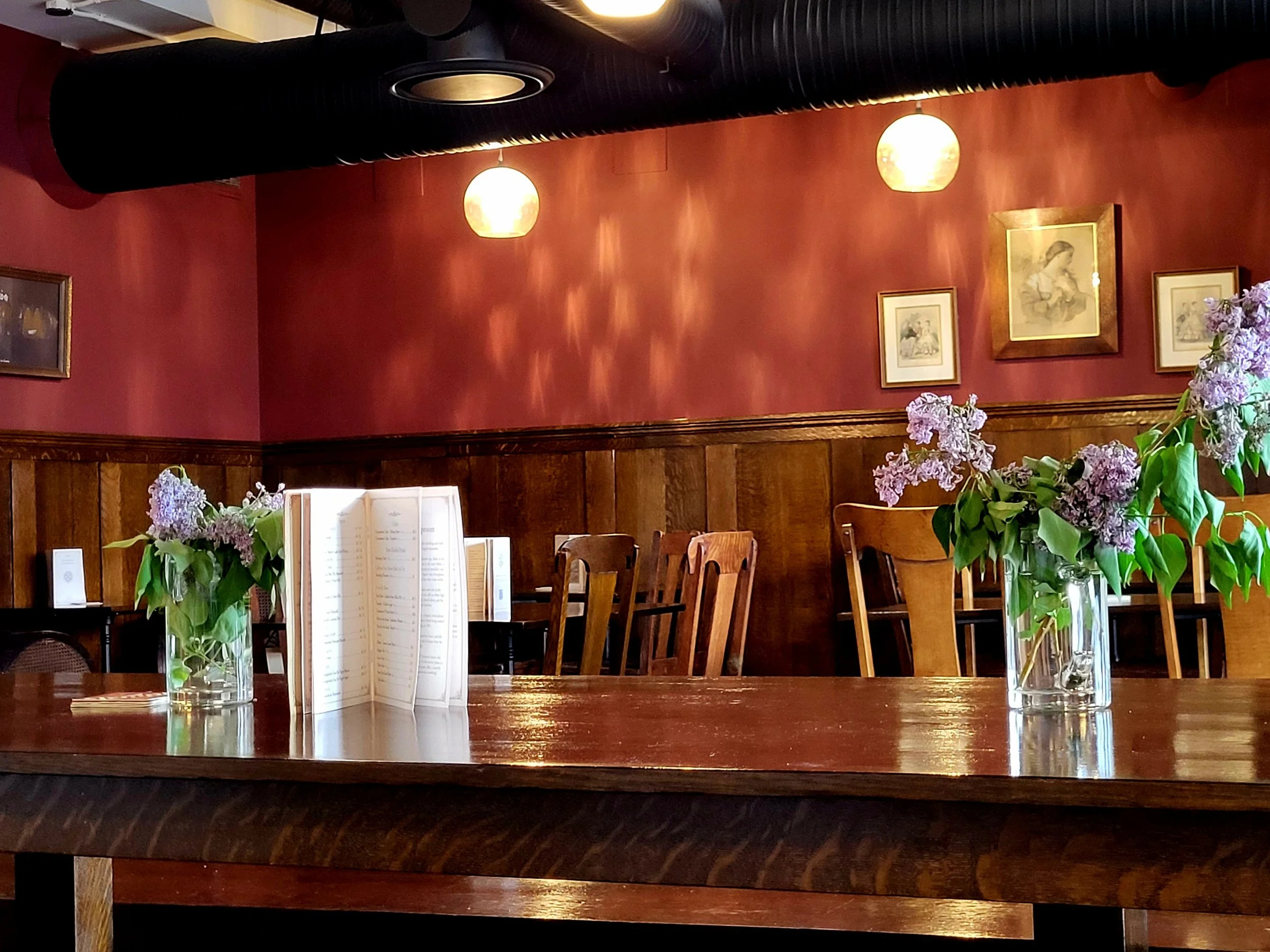 A wooden table with two glass vases filled with lilacs, a menu, and a wooden chair in a cozy restaurant with red walls, framed pictures, and hanging lights.