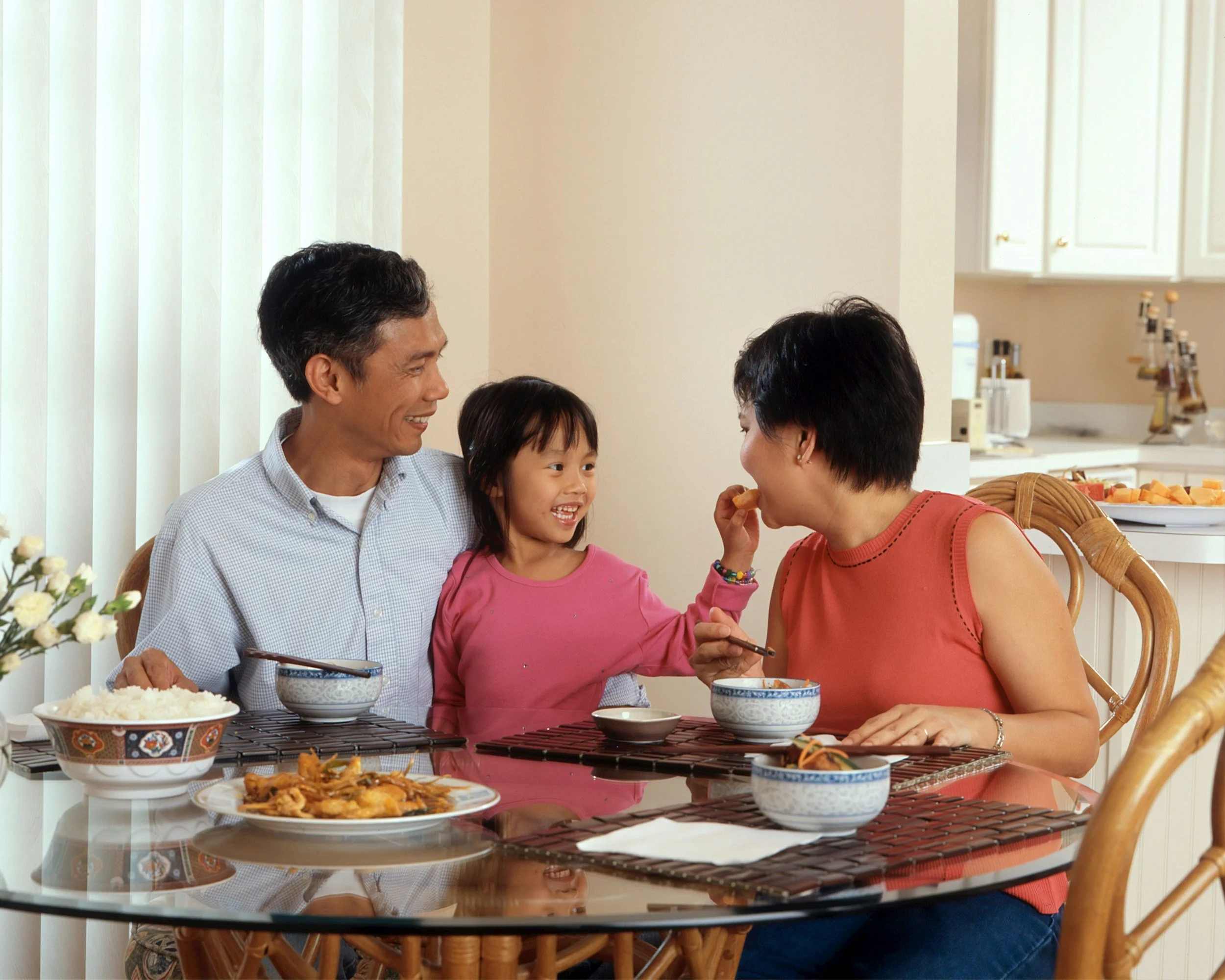 A family of three, with an older man, a young girl, and an adult woman, sit at a dining table sharing a meal. The girl is touching the woman's face affectionately, and everyone is smiling.