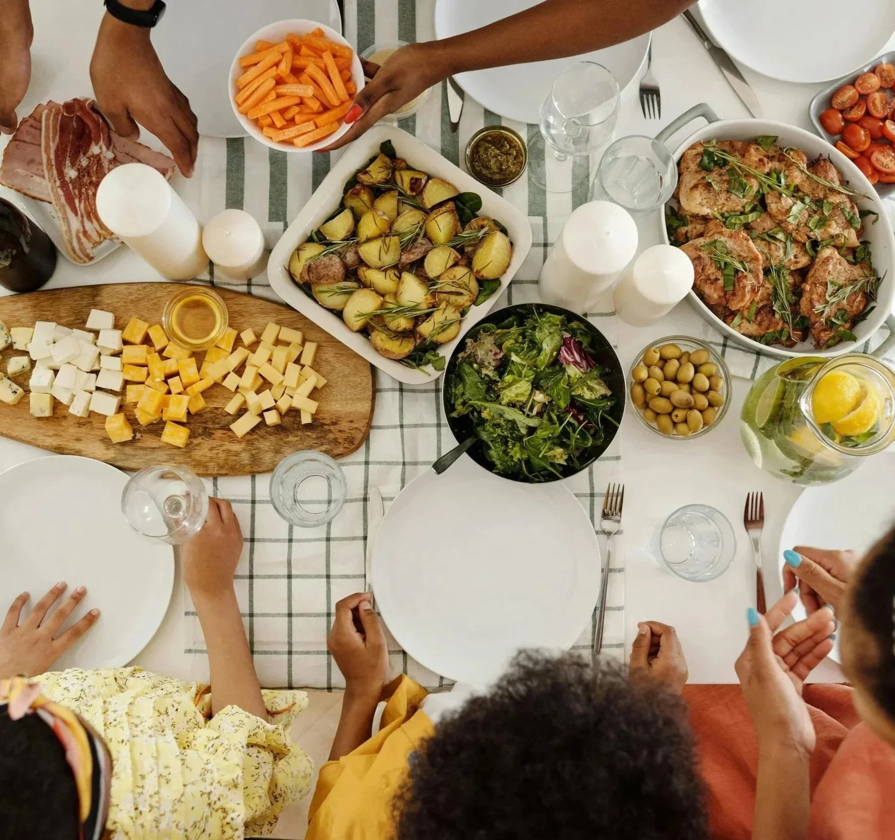 A family or group of people gathered around a dinner table with various foods including cheese, roasted potatoes, salad, chicken, carrots, cherry tomatoes, and drinks, as they prepare to eat.