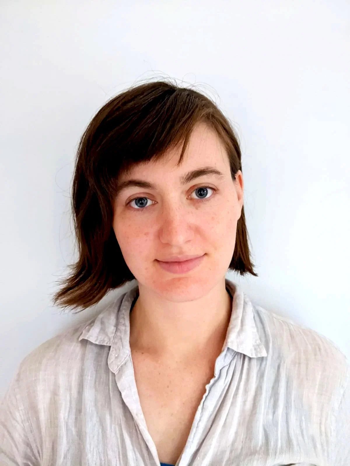 A young woman with short brown hair and blue eyes, wearing a light beige shirt, standing against a plain white wall.