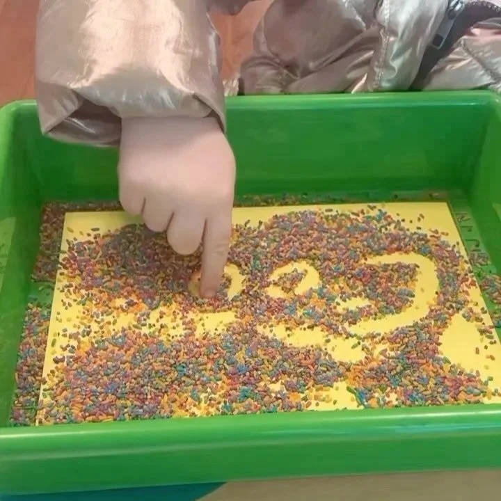 A person's hand, wearing a glove, pointing at a yellow surface covered with colorful sprinkles inside a green tray.