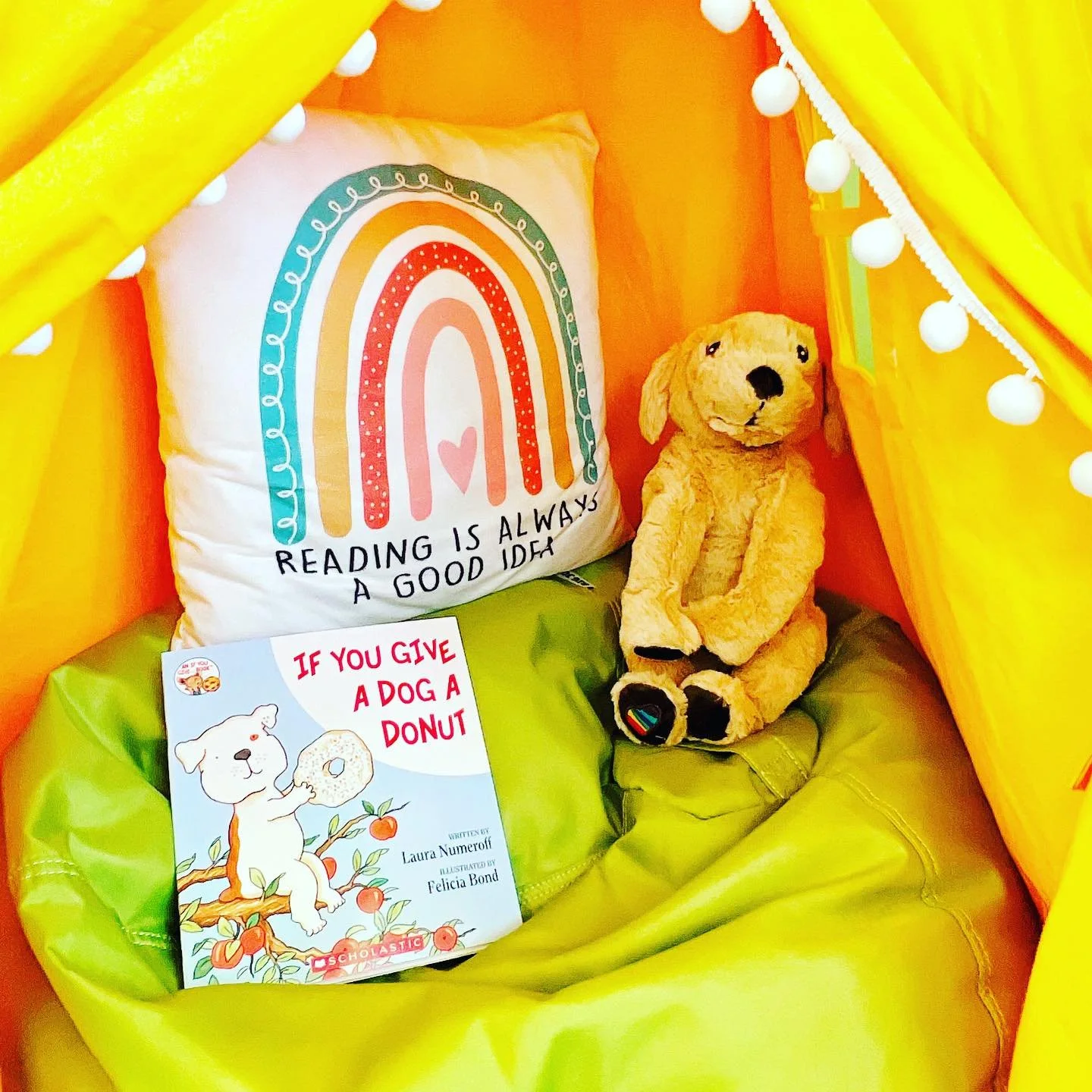 Colorful reading nook with a rainbow pillow featuring the quote 'Reading is always a good idea,' a plush dog toy, and a children's book titled 'If You Give a Dog a Donut' by Laura Numeroff, decorated with yellow curtains and string lights.