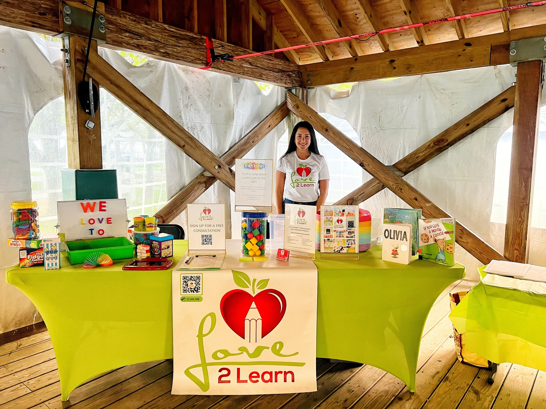 Remy Silvestre is standing behind a table display at a booth with a green tablecloth, showcasing children's educational materials and toys, with signs promoting free consultations and a QR code.