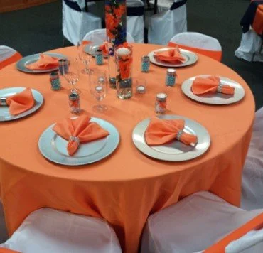 Round table with orange tablecloth, six white plates with orange napkins, small water glasses, and salt and pepper shakers, with a tall floral centerpiece in the middle.