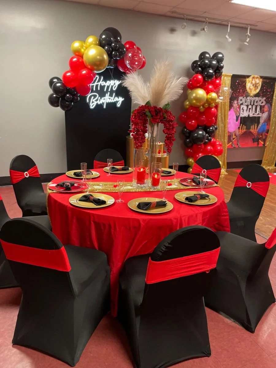 Round table with black chairs decorated with red ribbons, set for a celebration, with a large centerpiece of pampas grass and red flowers, and a backdrop with black, red, and gold balloons and a 'Happy Birthday' sign.