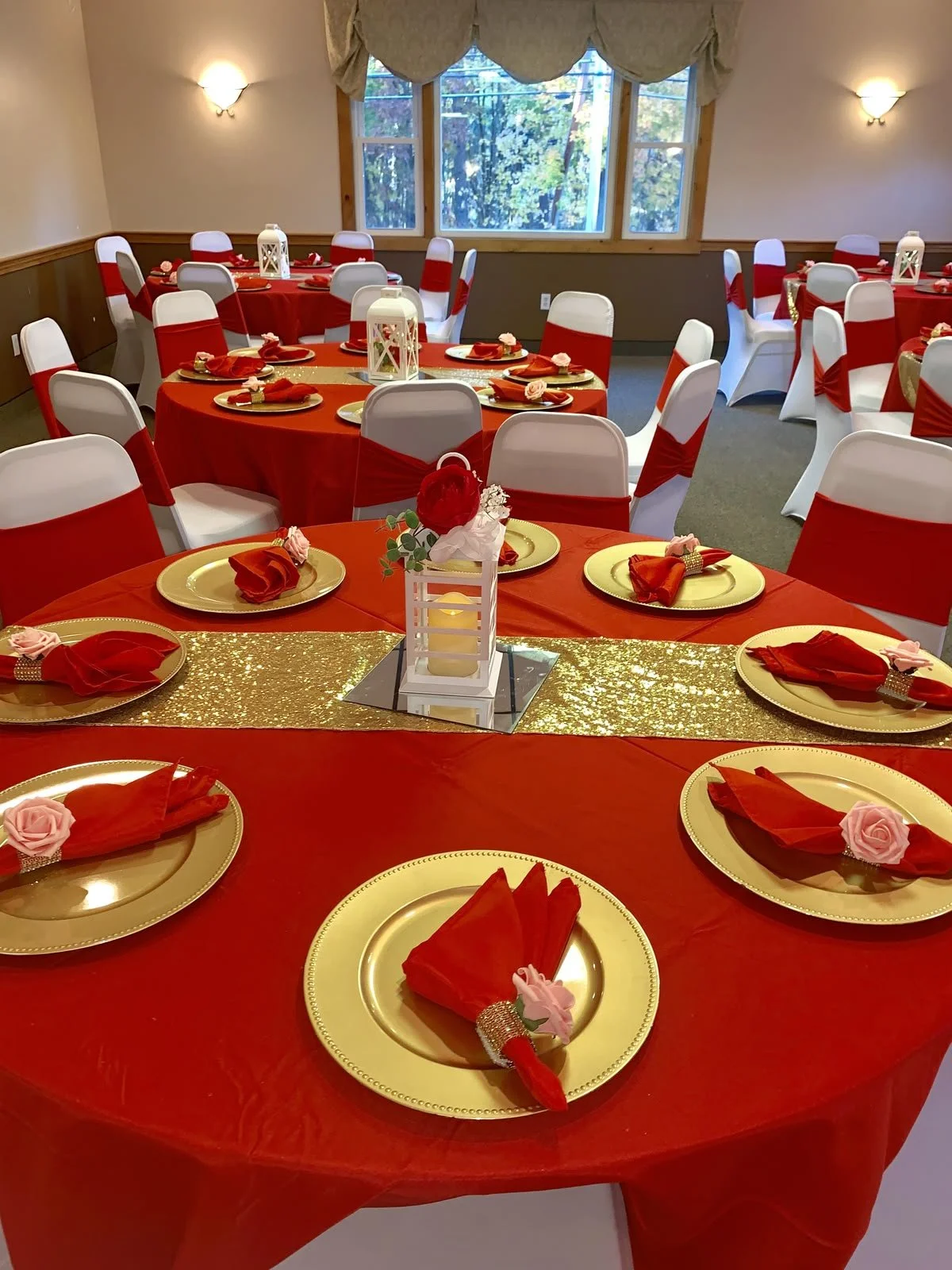 A decorated banquet room with tables draped in red tablecloths, set with gold plates, red napkins, and pink roses, featuring a window with a view of trees outside.