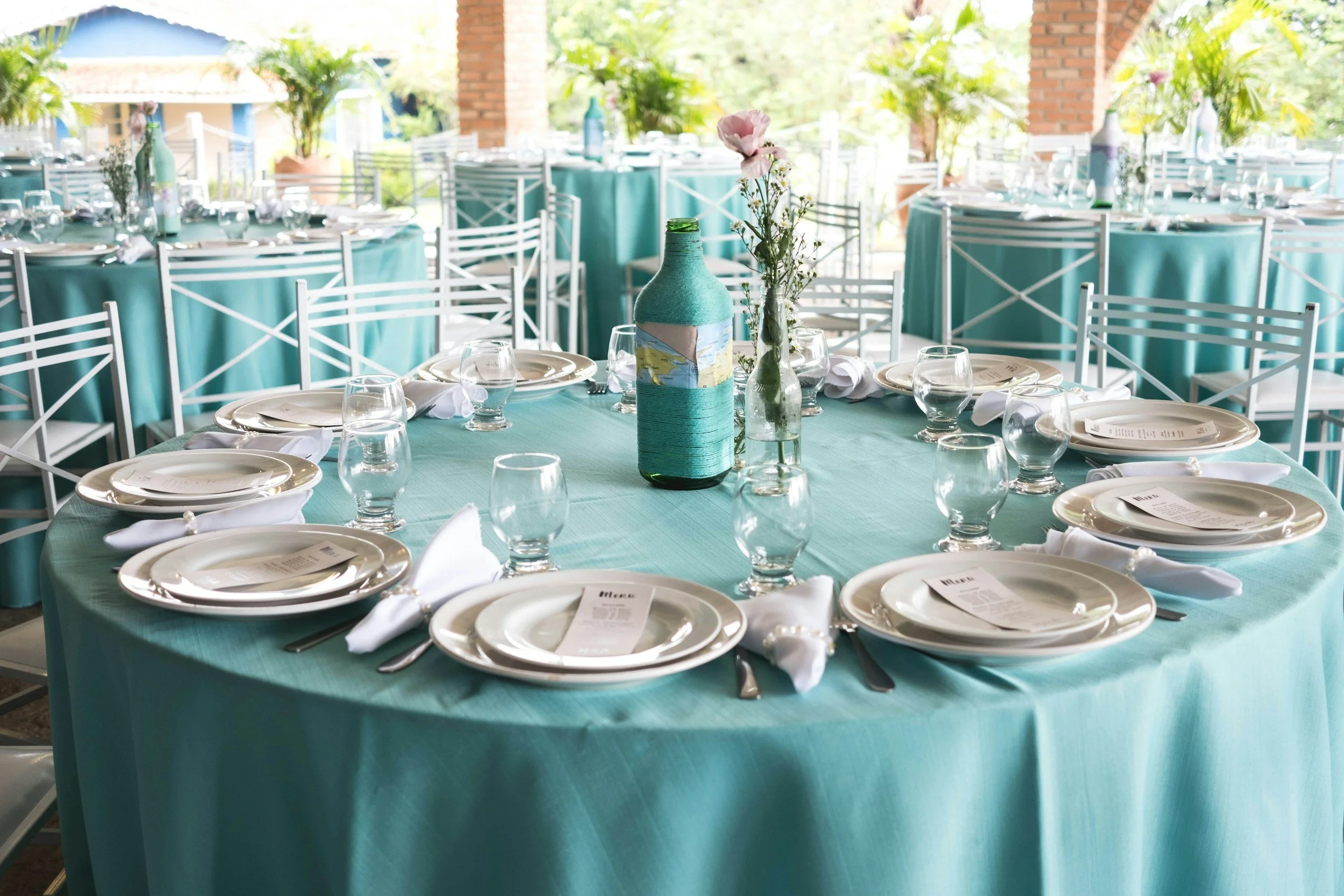 Round table set with white plates, silverware, water glasses, and white napkins, with a tall bottle covered in turquoise yarn and a small vase with pink and white flowers as centerpiece, in an outdoor covered patio area with turquoise tablecloths and white chairs.