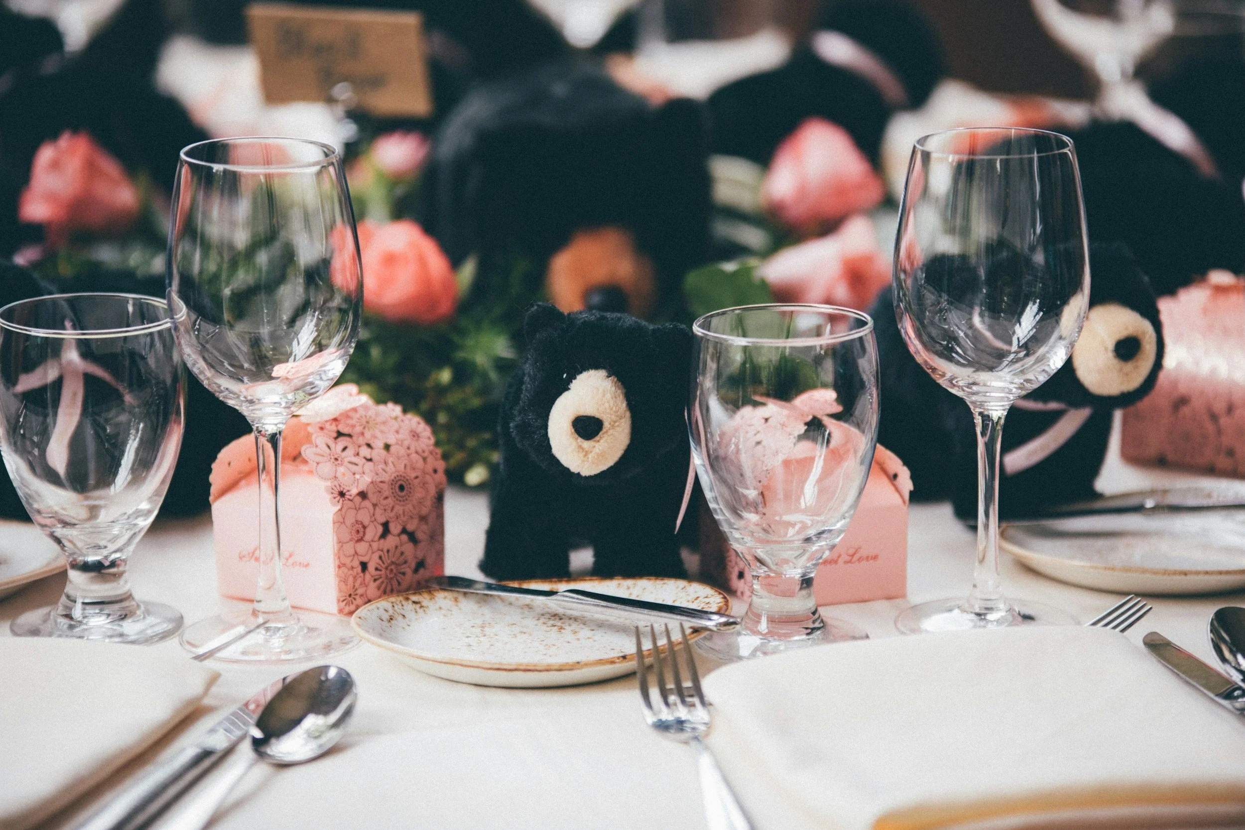 A table setting with three empty wine glasses, a small empty plate with a spoon, and pink gift boxes, surrounded by plush teddy bears and floral decorations.