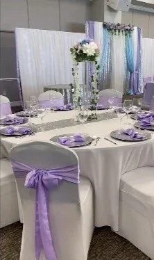 Elegant event table with white tablecloths, purple ribbons on chairs, and lavender-themed table settings, including napkins and floral centerpieces.