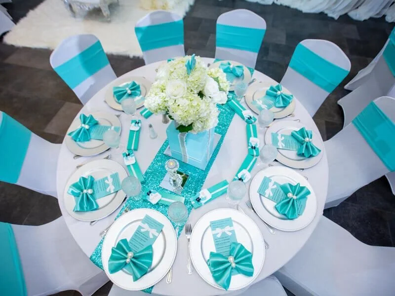 Round table decorated for a celebration with a white tablecloth and a turquoise glittery table runner. White plates each with a turquoise paper napkin folded into a bow, pearl accents, and cutlery. A centerpiece of white flowers in a turquoise vase. The chairs are covered in white with turquoise sashes.