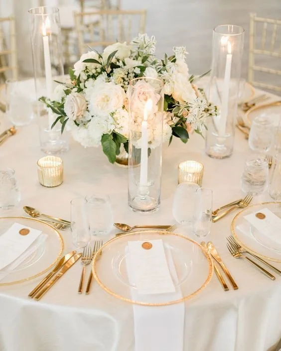 Elegant wedding reception table with white tablecloth, gold-rimmed dinnerware, gold flatware, and white napkins. Centerpiece features a tall glass vase with white and blush flowers, surrounded by tall glass candle holders with lit white candles, and small gold votive candles.