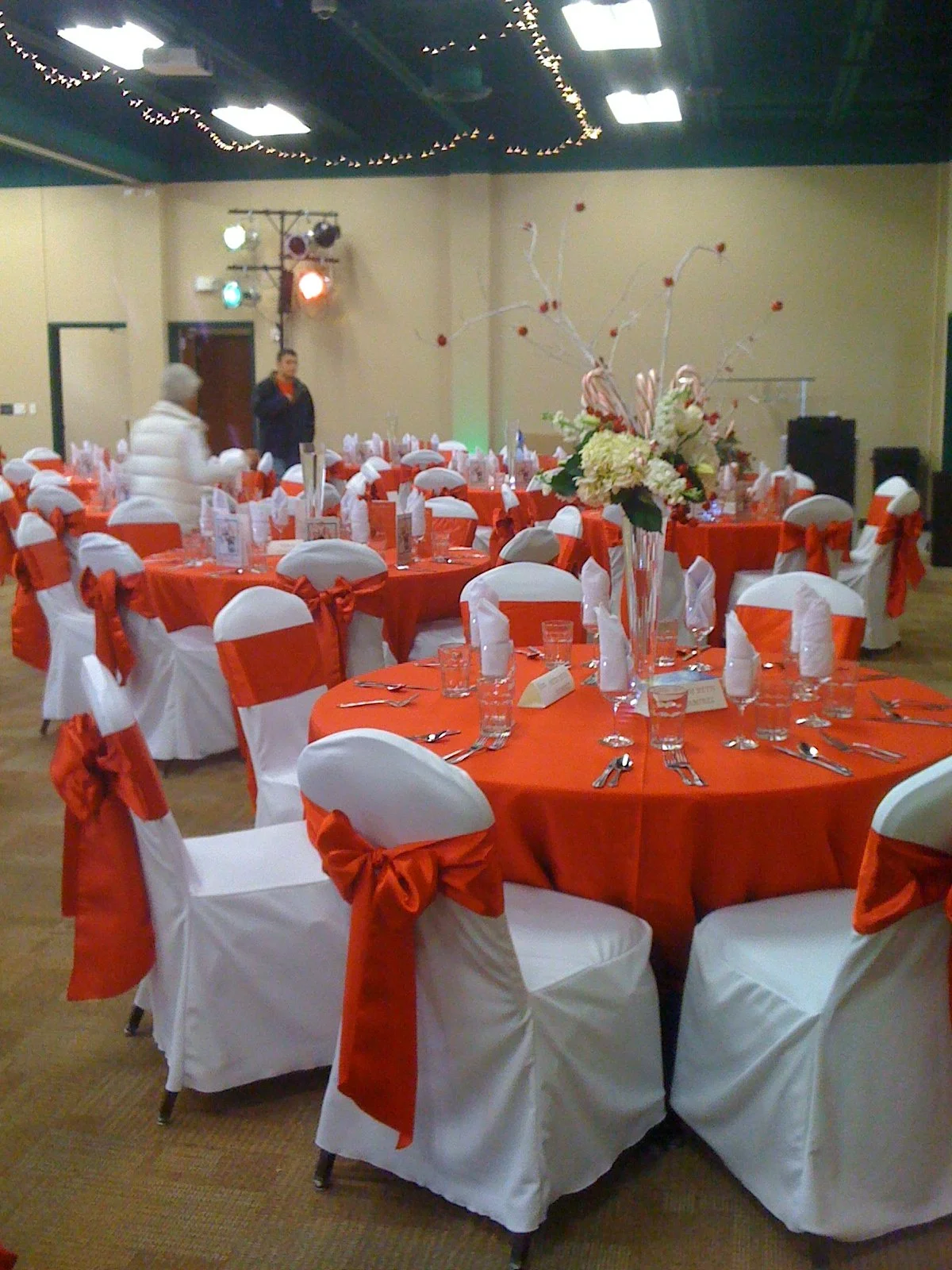 Event hall decorated with round tables covered in orange tablecloths and white chairs with orange sashes, centerpieces with flowers, and place settings for a celebration or banquet.