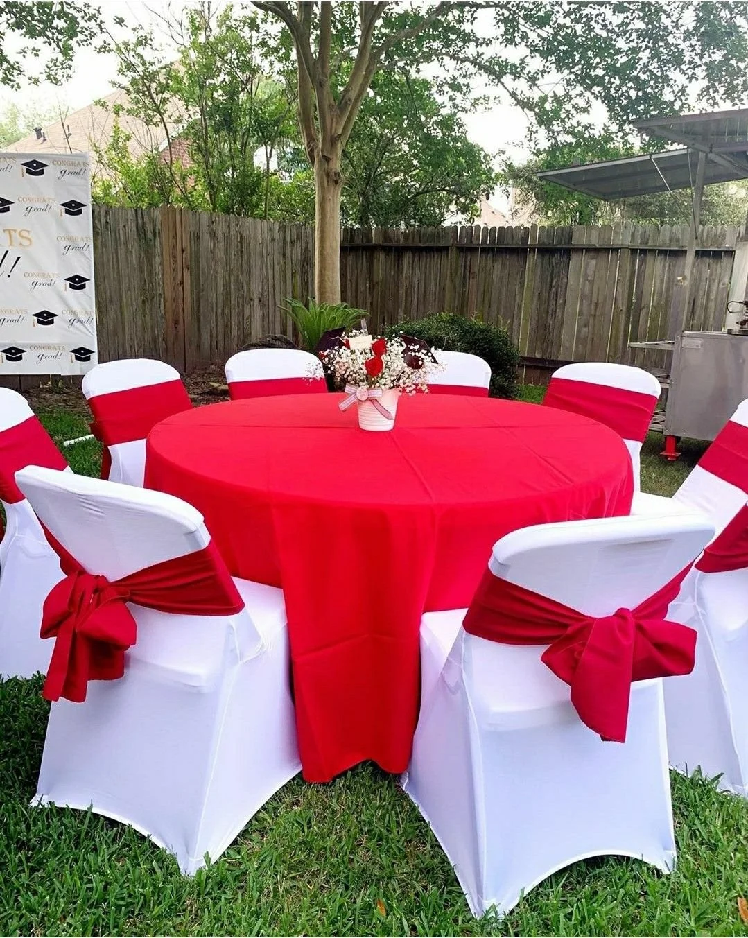 Outdoor party table with a red tablecloth and white chairs decorated with red sashes, set in a backyard with trees, a wooden fence, and a congratulatory sign.