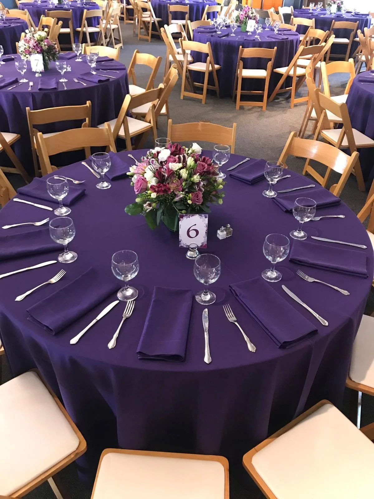 Round banquet table set with purple tablecloth, floral centerpiece, purple napkins, glassware, and silverware, in a decorated event hall.