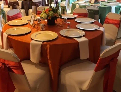 Round banquet table set with orange tablecloth, white napkins, plates, and a floral centerpiece, surrounded by chairs with colorful sashes, at an event or celebration.