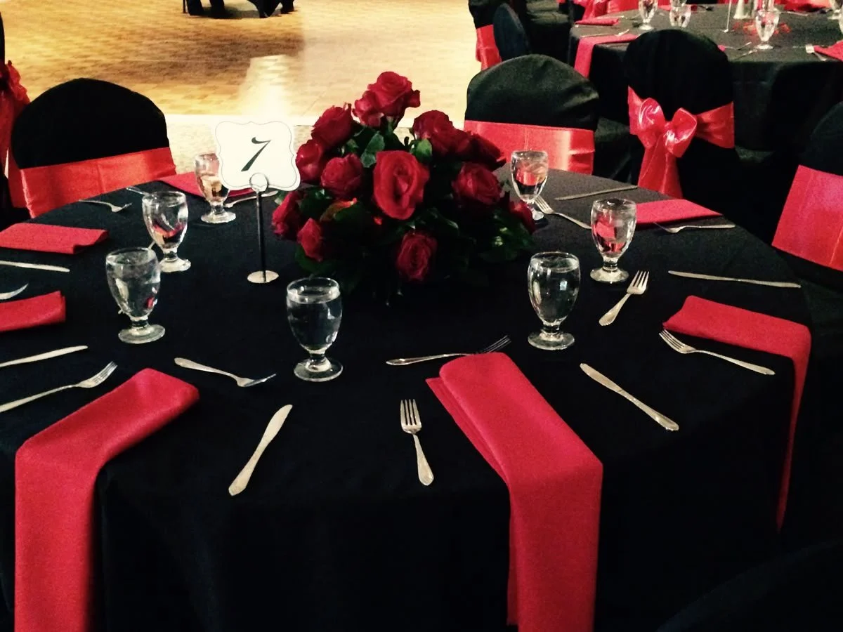 Formal event table setting with red napkins and a centerpiece of red roses, black tablecloth, black chairs with red sashes, and place settings including forks, knives, and glasses.