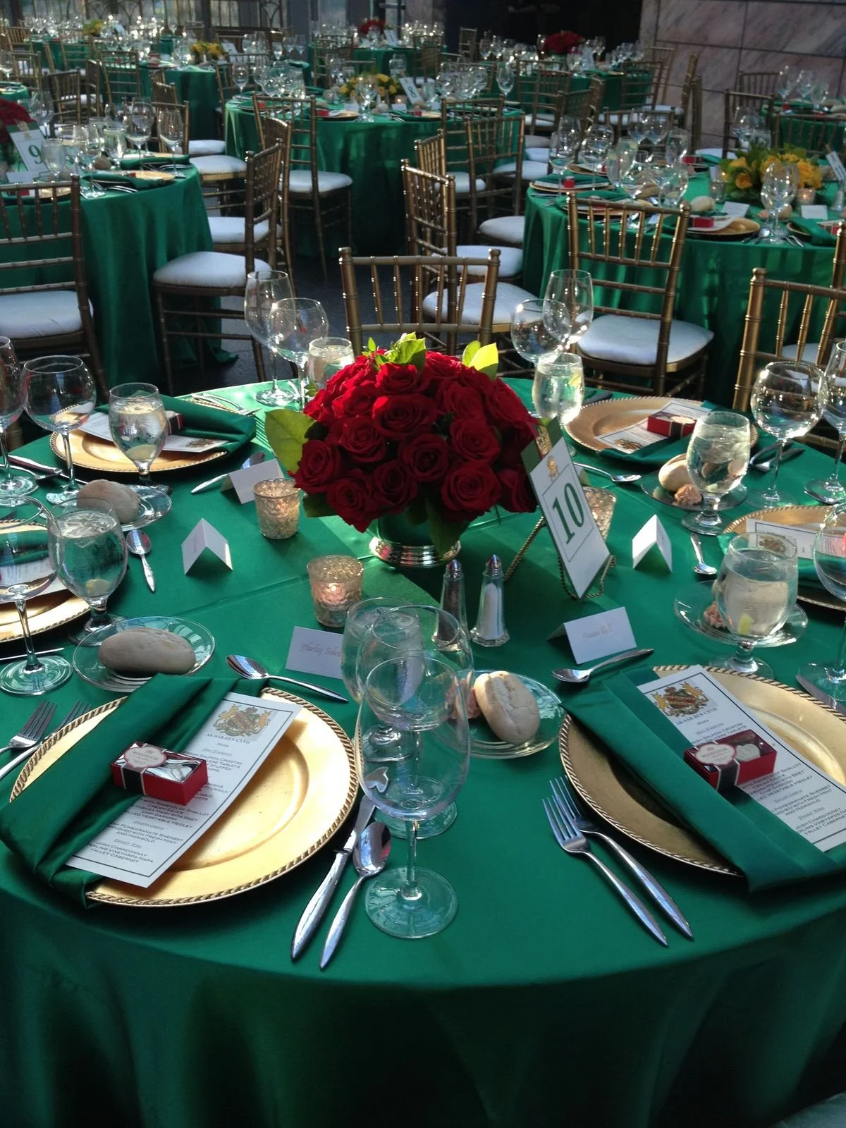A banquet table decorated with green tablecloths, gold chargers, green napkins, wine glasses, and place settings, with a centerpiece of red roses and multiple small candles.
