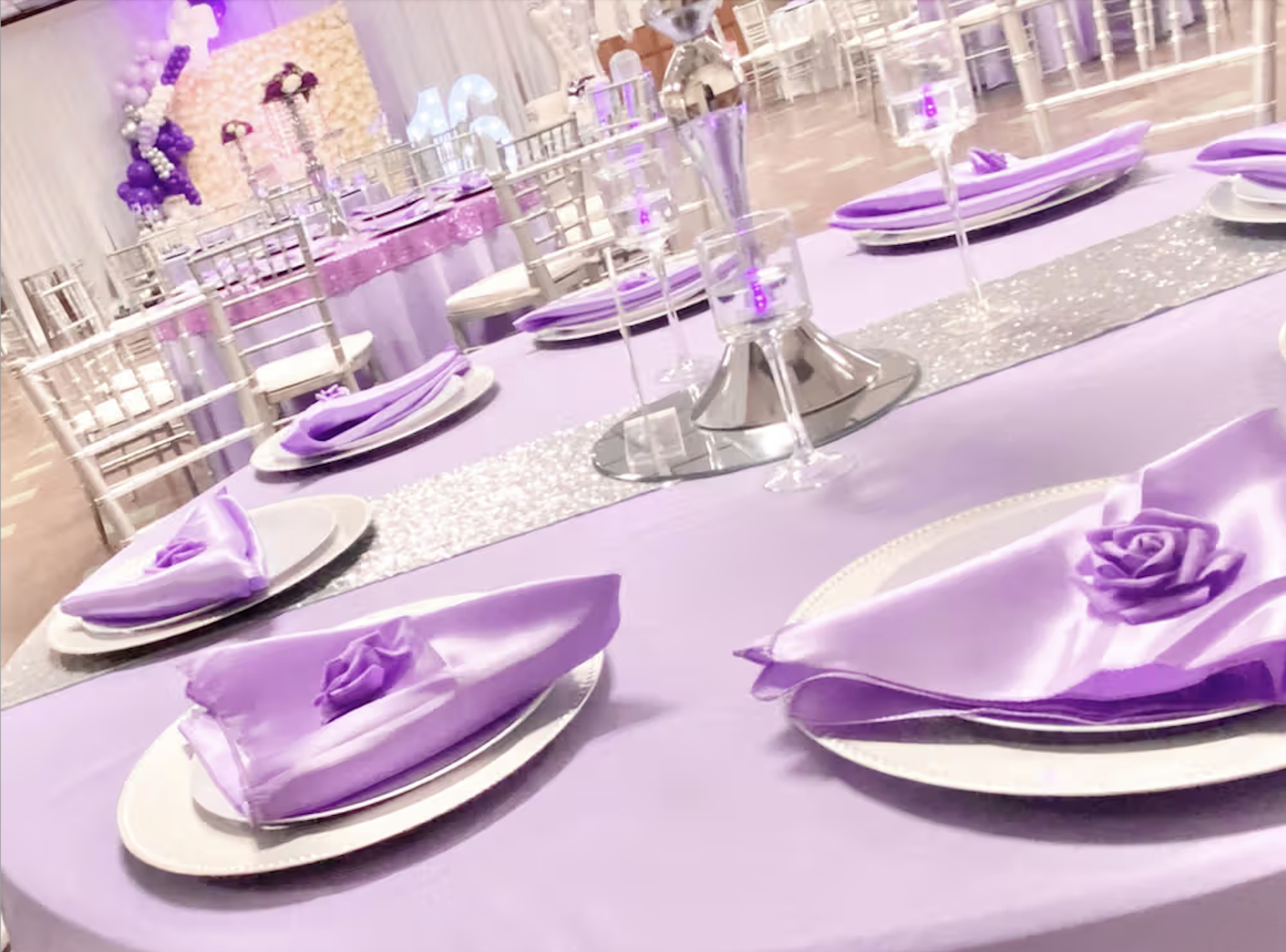 Elegant event table setup with purple accents, decorated plates with purple napkins folded in a rose shape, tall crystal candle holders, and a background with purple and white balloons and flowers.