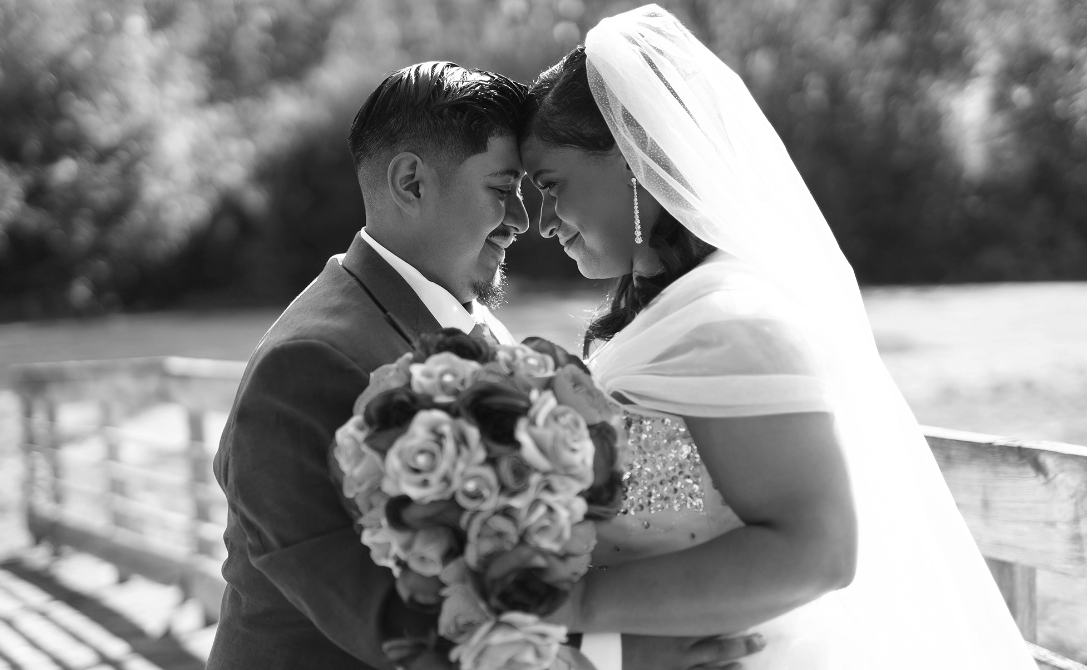 A bride and groom with foreheads touching, smiling, on a wooden bridge, holding a bouquet of flowers, black and white photo.