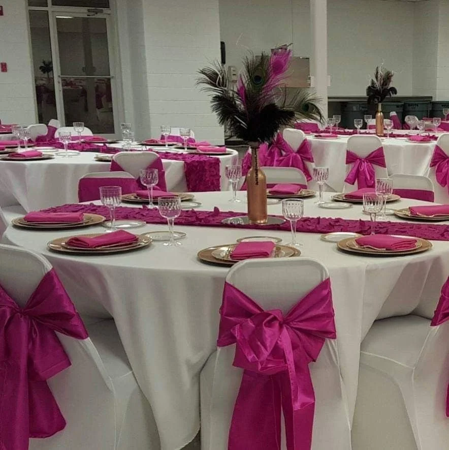 Clothed banquet tables with white tablecloths and pink accents, set with plates, crystal glasses, and pink napkins, decorated with centerpieces of pink peacock feathers in gold vases, in a decorated event hall.