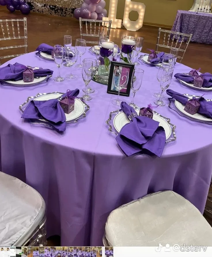 Round table decorated for a celebration with a purple tablecloth, purple napkins, and small purple gift boxes at each place setting. The table has glassware and a centerpiece with candles and greenery. In the background, there are balloon decorations, large illuminated letters, and another decorated table.