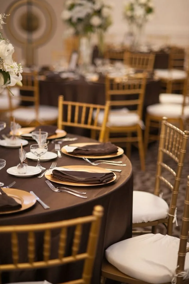 Elegantly set round table with gold plates, dark napkins, clear drinking glasses, and silverware, prepared for a formal event in a decorated venue with floral centerpieces and chairs with white cushions.