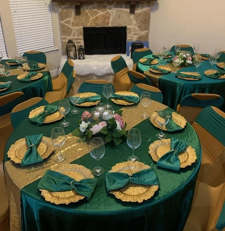 Round tables decorated with green tablecloths, gold placemats, and teal napkins tied with gold rings, set with wine glasses and centerpieces of pink and white flowers. Chairs draped in yellow and green covers surround the tables. A stone fireplace with lanterns on the hearth is in the background.